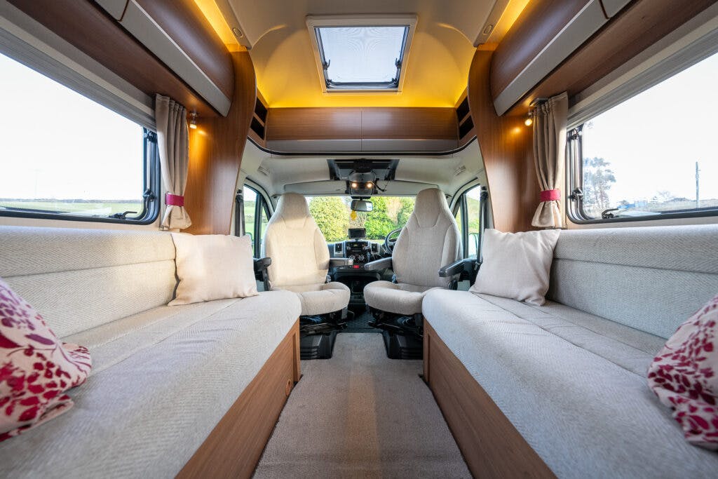 Inside the 2014 Auto-Trail Imala 715 Lowline, you'll find two beige sofas on either side, adorned with light-colored cushions. A central aisle leads to the driver and passenger seats, while a skylight and windows flood the space with natural light.