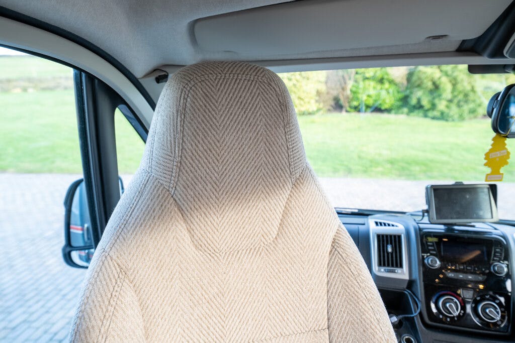 A driver's seat with a gray herringbone-patterned cover in the 2014 Auto-Trail Imala 715 Lowline's interior. Visible in the background are a steering wheel, dashboard with various controls, air freshener, GPS device, and a side mirror showing a view of a grassy outdoor area.