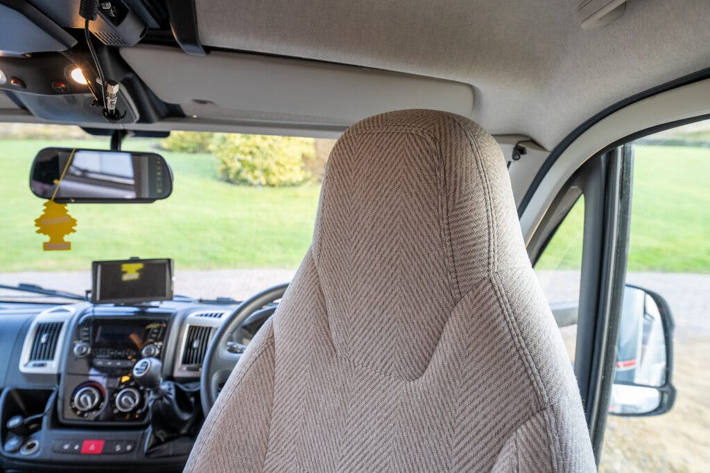 Interior view of a 2014 Auto-Trail Imala 715 Lowline focusing on the front driver's seat and dashboard. The seat is covered in a patterned fabric. The dashboard features various controls, a GPS device, and a rearview mirror with an air freshener hanging from it. The passenger side door is open.