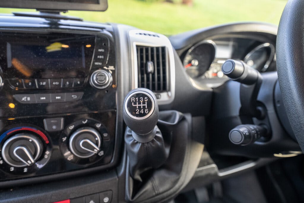 The image shows the interior of a 2014 Auto-Trail Imala 715 Lowline with a manual transmission. The focus is on the gear shift knob, which displays six gears and reverse. The dashboard includes control knobs and buttons, an air vent, and part of the steering wheel is visible.