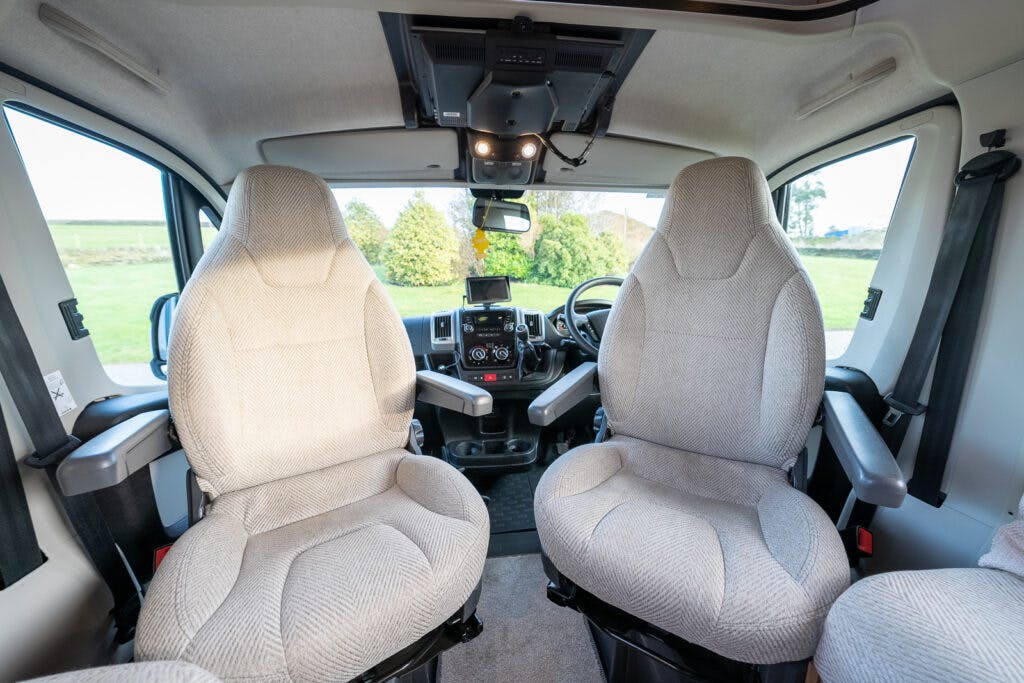 The interior of the 2014 Auto-Trail Imala 715 Lowline features two front seats facing the camera, upholstered in light gray fabric. The windshield and side windows offer a view of greenery outside. The dashboard and various controls are visible between the seats.