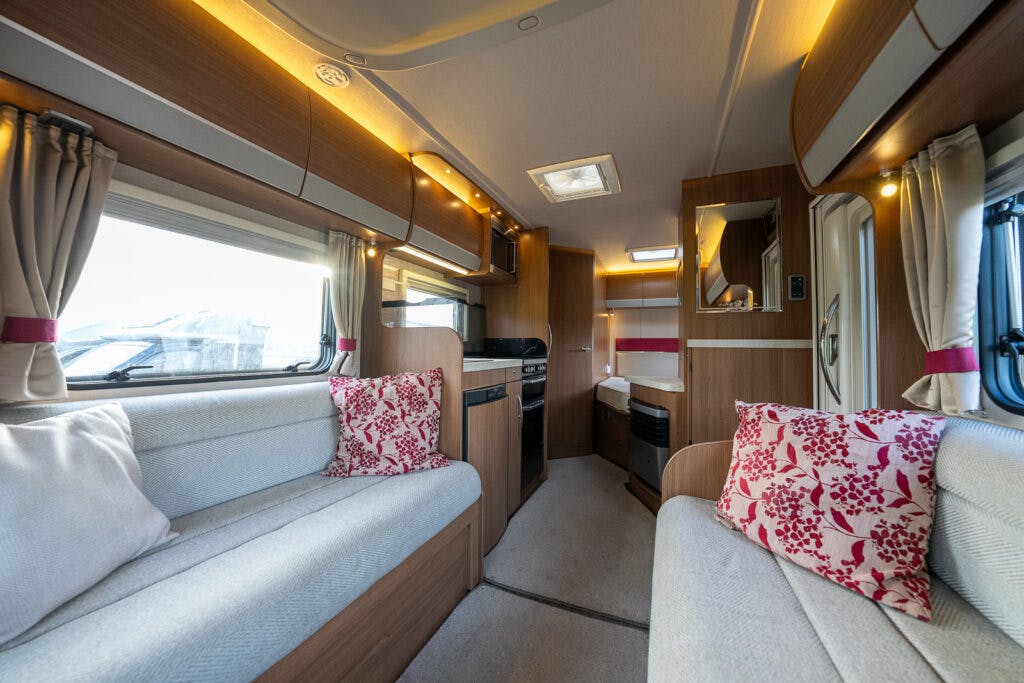 Interior of the 2014 Auto-Trail Imala 715 Lowline showcases two long beige sofas with pink and white floral cushions on either side. The space includes wooden cabinetry, a small kitchen area with an oven, and a door leading to a backroom. Windows are adorned with beige curtains.