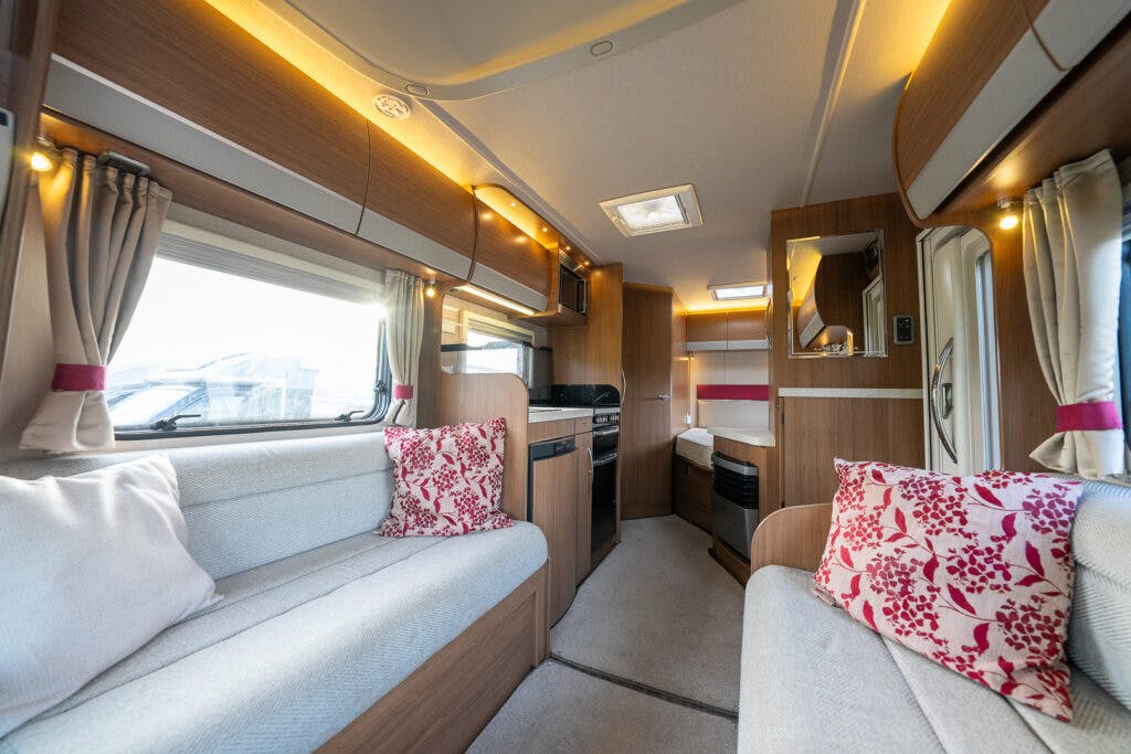 The interior of the 2014 Auto-Trail Imala 715 Lowline RV is shown, featuring light wood finishes and beige upholstered seating with red and white patterned cushions. The RV contains a kitchenette with a stove, storage cabinets, overhead lighting, and windows with curtains.