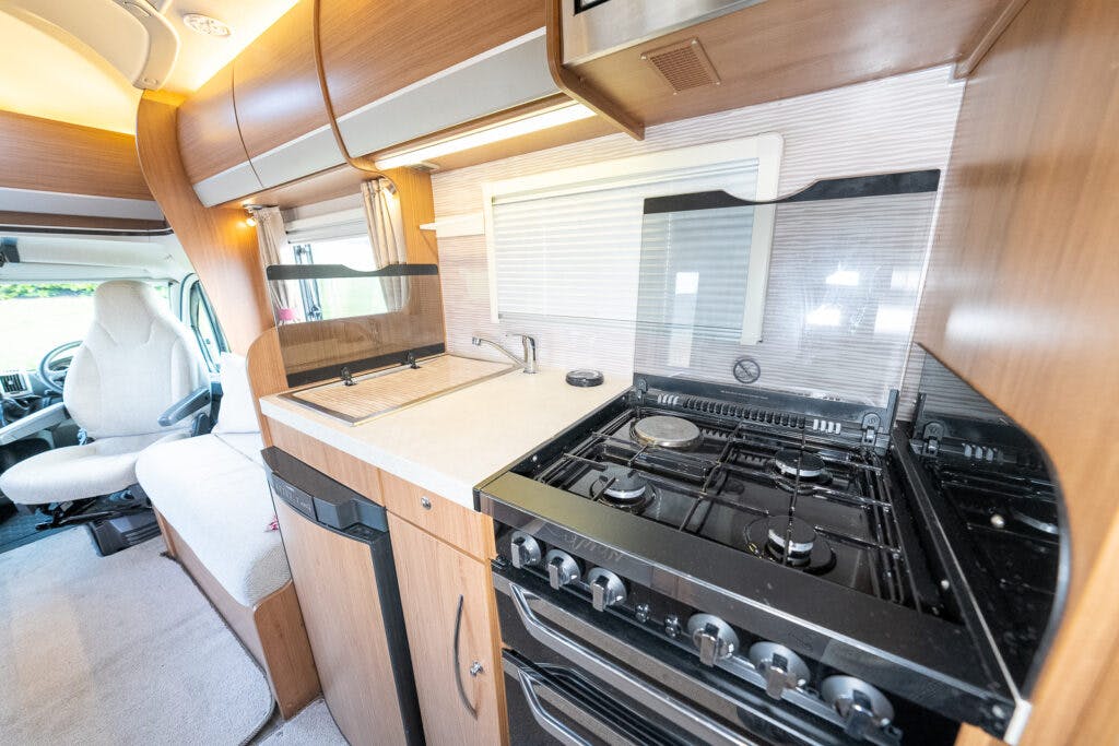 A 2014 Auto-Trail Imala 715 Lowline motorhome kitchen features a stove with multiple burners, an oven below, and a sink next to it. The area includes wooden cabinetry, a small countertop, and a window above the sink. An upholstered driver's seat and a cockpit area are visible to the left.