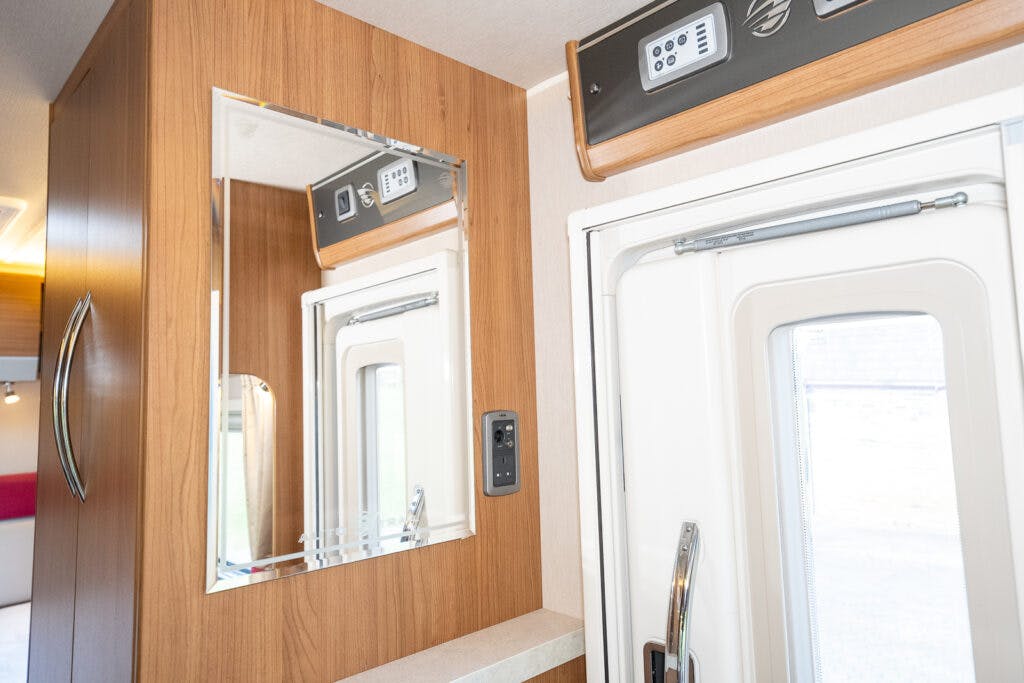 The image shows an interior corner of a 2014 Auto-Trail Imala 715 Lowline RV. Visible are a wooden wall with a mounted mirror, part of an electronic control panel above the door frame, and an open white door with a handle.