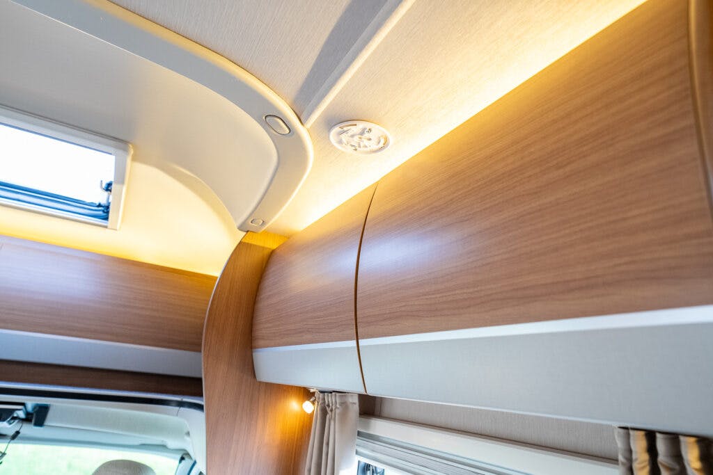 A close-up view of the interior ceiling of a 2014 Auto-Trail Imala 715 Lowline, featuring built-in wood grain storage cabinets with integrated lighting along the top. There is also a round ceiling light fixture and a small window on the left side.