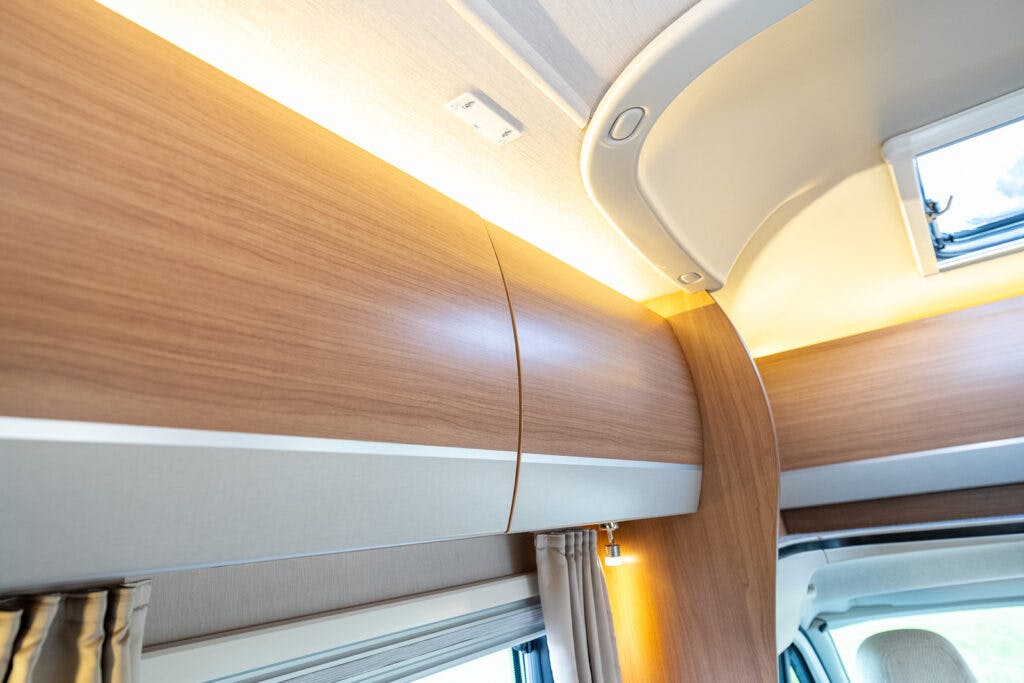 Interior view of a 2014 Auto-Trail Imala 715 Lowline RV with wooden cabinets and soft lighting. The curved design of the overhead storage and the light-colored ceiling create a cozy and modern atmosphere. A small window is partially visible in the top right corner.
