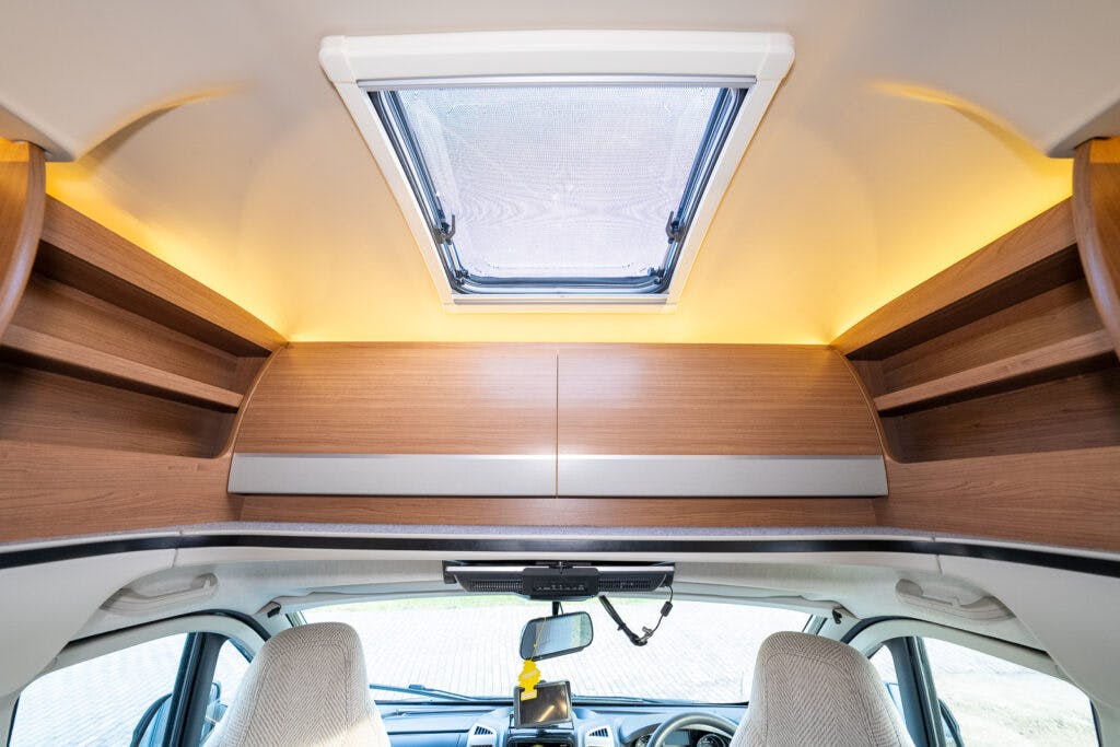 Interior view of a 2014 Auto-Trail Imala 715 Lowline motorhome showing two overhead storage compartments with wooden finishes, a skylight window, and the front dashboard area with seats. The image highlights the practical use of space and storage options within this well-designed motorhome.