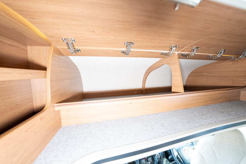 The image shows an open storage compartment in what appears to be a 2014 Auto-Trail Imala 715 Lowline, likely a camper van or RV. The compartment features wooden panels with multiple hinge-supported doors opened upward. Inside, there are shelves and a carpeted bottom surface, with car controls faintly visible below.