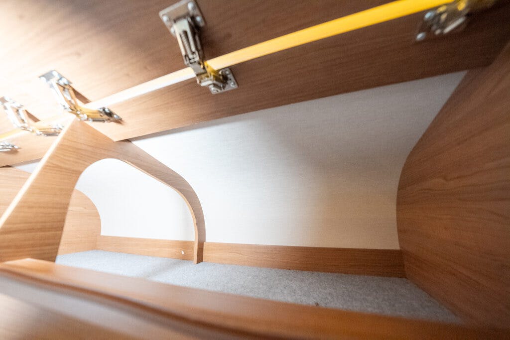 Interior view of an empty wooden storage compartment with light-colored lining and metal hinges. The space includes an arch-shaped opening, reminiscent of the practical design seen in a 2014 Auto-Trail Imala 715 Lowline, likely serving as under-bed storage with a lifting mechanism.