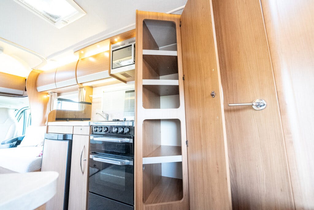 Interior of the 2014 Auto-Trail Imala 715 Lowline camper van featuring a compact kitchen. The kitchen includes a stove, small oven, overhead microwave, and open wooden shelving units. The design shows a mix of metal and wood finishes, with ambient lighting above the stove.