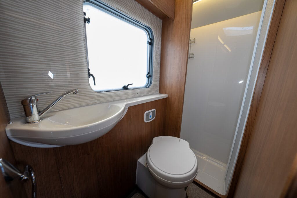 This image shows the small bathroom of a 2014 Auto-Trail Imala 715 Lowline recreational vehicle. It features a white sink with a single-handle faucet, a white toilet, and a shower area with translucent doors. A window above the sink has a shade partially pulled down. Walls and cabinets are wooden.