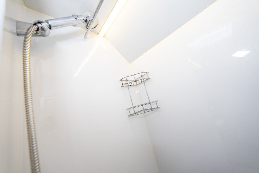 A white shower stall in the 2014 Auto-Trail Imala 715 Lowline features a metal adjustable showerhead with a flexible hose and a silver corner shelf for toiletries. The lighting is bright, and the stall has clean, smooth surfaces.