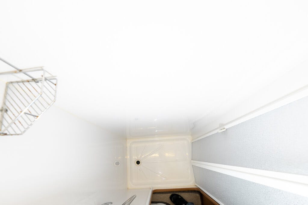 A small, white shower stall with a metal corner shelf, reminiscent of the compact design found in a 2014 Auto-Trail Imala 715 Lowline. The photo captures an overhead view of the space, showing the square floor pan with a central drain. Part of a black shoe and a floor mat are visible outside the shower.