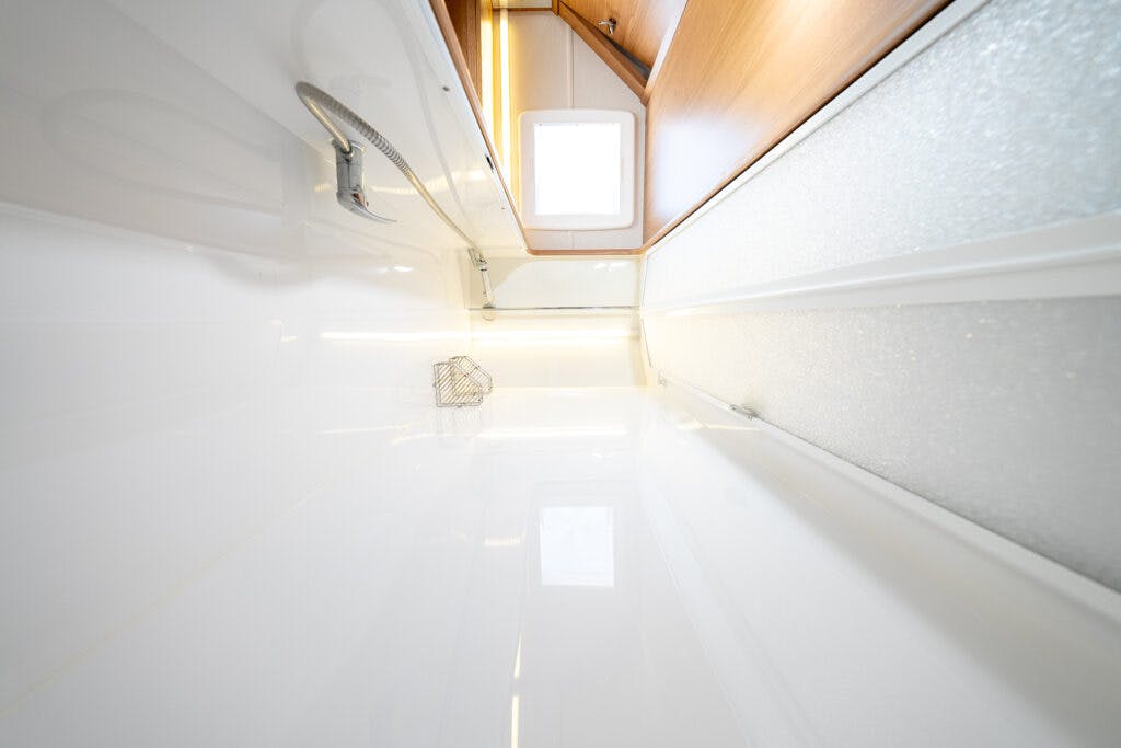 The image shows a narrow, enclosed space within the 2014 Auto-Trail Imala 715 Lowline, featuring white, glossy walls and a small frosted window. A metallic wire basket is mounted on one wall, complemented by overhead wooden cabinets. Bright lighting illuminates the area.