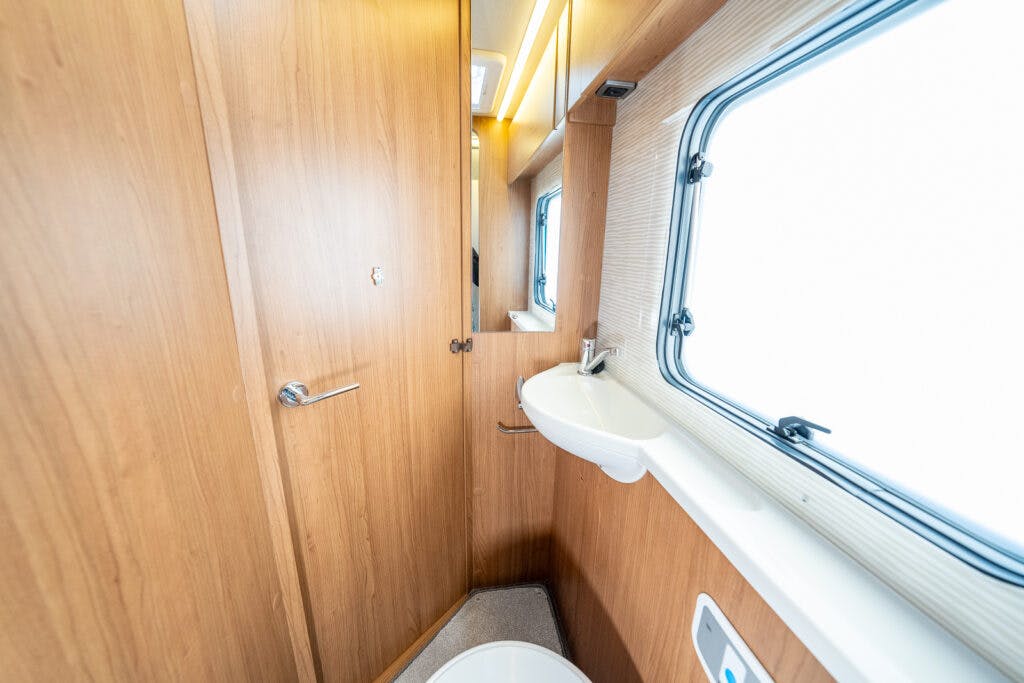 The image shows a compact bathroom inside a 2014 Auto-Trail Imala 715 Lowline, featuring a small white sink and a mirror mounted on a wood-paneled wall. A window with a closed blind is above the sink, and a door with a handle is partially visible.