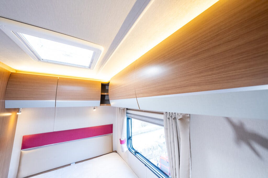 Interior view of the 2014 Auto-Trail Imala 715 Lowline camper van, showcasing wooden cabinetry and a bed with a light beige cushion and pink headboard. Natural light streams through a side window with curtains and a roof vent, while lighting under the cabinets creates a warm ambiance.