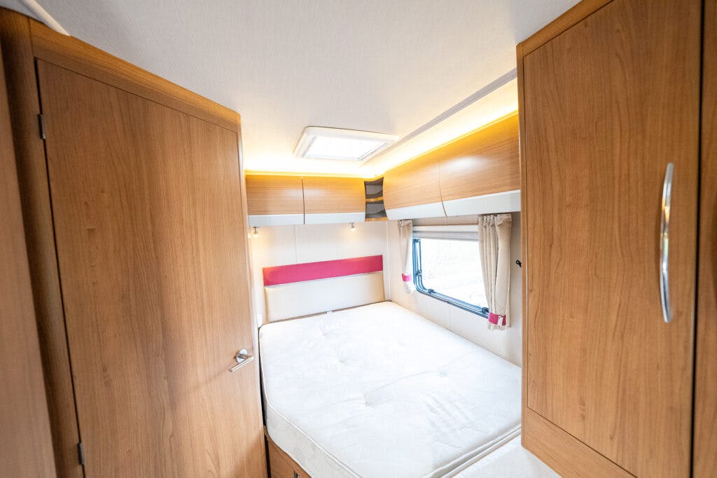 Interior of the 2014 Auto-Trail Imala 715 Lowline camper van bedroom featuring a double bed with a white mattress, a small window with curtains, and wooden cabinetry on both walls and ceiling. The room is well-lit with ceiling and under-cabinet lighting.