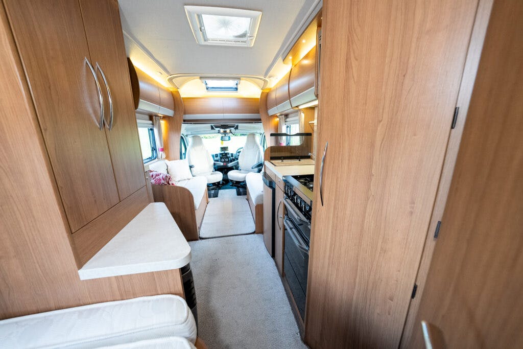 The photo shows the interior of a 2014 Auto-Trail Imala 715 Lowline motorhome. The view is along a narrow corridor leading to the driver's area, with wooden cabinetry on each side, a small kitchen with an oven and stove to the right, and seating at the far end.