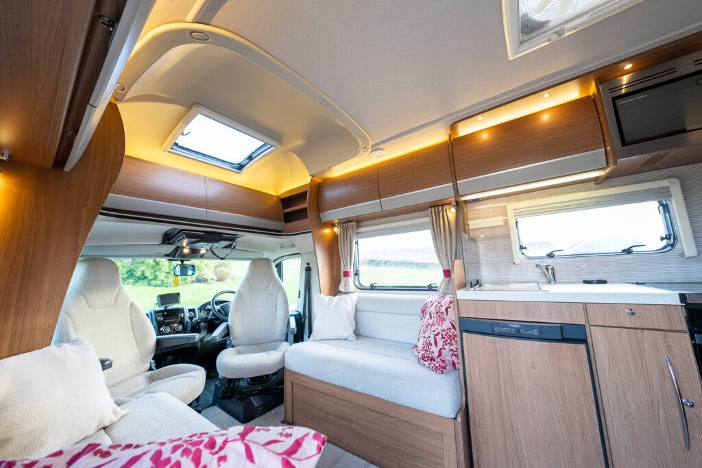 Interior of the 2014 Auto-Trail Imala 715 Lowline campervan featuring a seating area with light gray cushions and white pillows, a kitchenette with a counter and cabinets, and the driver's area visible at the front. The space is well-lit with natural light from windows and ceiling lights.