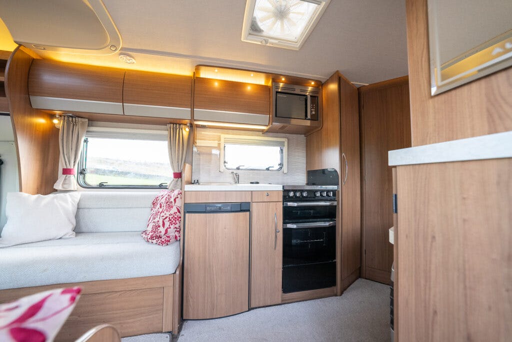 The interior of the 2014 Auto-Trail Imala 715 Lowline camper van features a kitchenette with wooden cabinets, a window, a sink, stove, oven, and microwave. Adjacent is a seating area with beige cushions and a red patterned blanket, set against a wall with another window.
