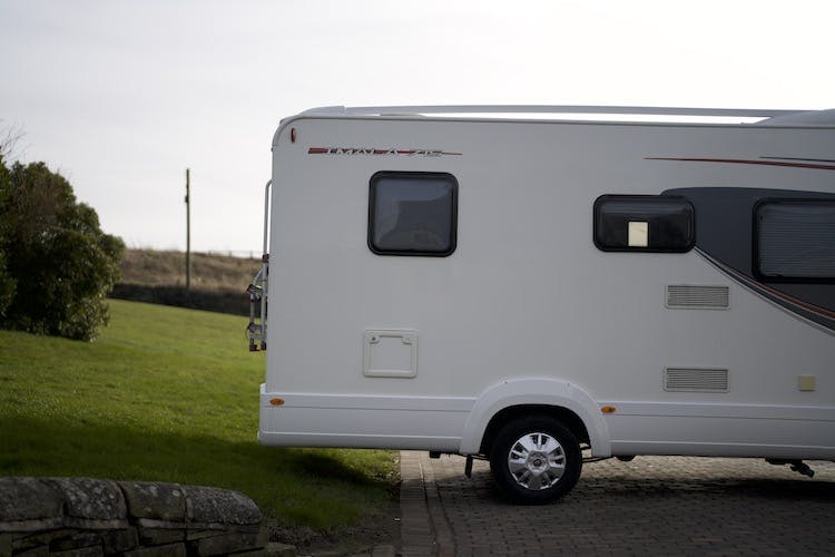 A 2014 Auto-Trail Imala 715 Lowline motorhome is parked on a paved driveway next to a grassy area. Part of a stone wall is visible in the bottom left, with a power line and grassy field in the background. The image is taken during the daytime.