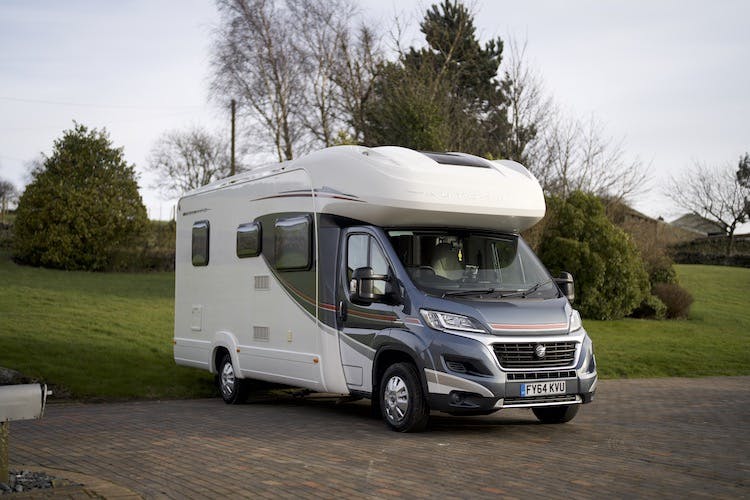 A modern 2014 Auto-Trail Imala 715 Lowline motorhome is parked on a brick driveway, set against a backdrop of lush green grass, trees, and shrubs. The sleek vehicle, white with gray and red accents, proudly displays its registration plate "FY64 KXU.