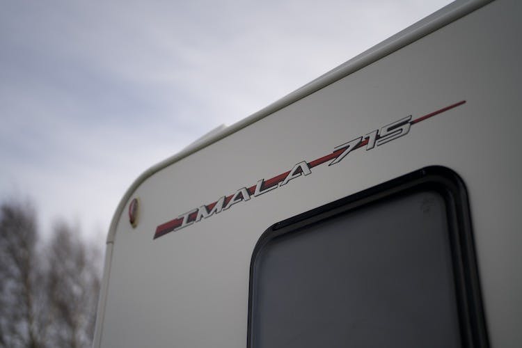 Close-up of the side of a 2014 Auto-Trail Imala 715 Lowline in white, showcasing "IMALA 715" in red and black next to a rectangular window. The sky is cloudy, and the tops of leafless trees are visible in the background.