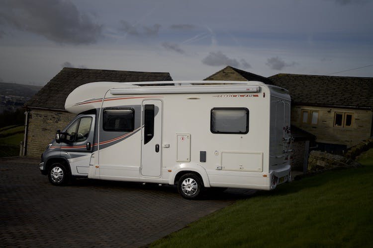 A 2014 Auto-Trail Imala 715 Lowline motorhome is parked on a slightly inclined driveway next to a stone building with a sloped roof. The sky is overcast, and the grass around the area is green. The motorhome has various windows and markings along its side.