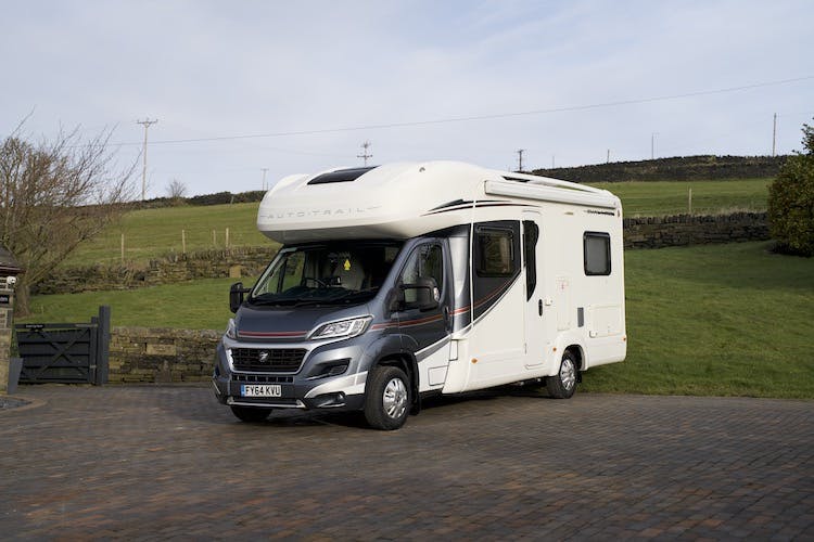 A 2014 Auto-Trail Imala 715 Lowline white motorhome parked on a brick driveway. The vehicle has a sleek design with a high roof, tinted windows, and various compartments. In the background, there is a stone fence, a grassy hill, and an overcast sky.