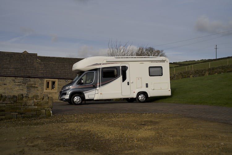 A white 2014 Auto-Trail Imala 715 Lowline motorhome is parked on a paved driveway next to a stone building. The background includes a grassy area and a stone wall, with a clear sky overhead. The motorhome has stripes on its side and a large front cab.