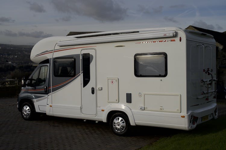 A 2014 Auto-Trail Imala 715 Lowline motorhome is parked on a brick driveway. It features decorative stripes along its side and has a rear bike rack. The motorhome is situated in front of a landscape under a clouded sky.