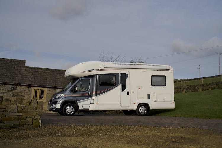 A white 2014 Auto-Trail Imala 715 Lowline motorhome is parked on a paved driveway next to a stone wall and building. It has black and red detailing and is situated in a rural area with green grass and an overcast sky.
