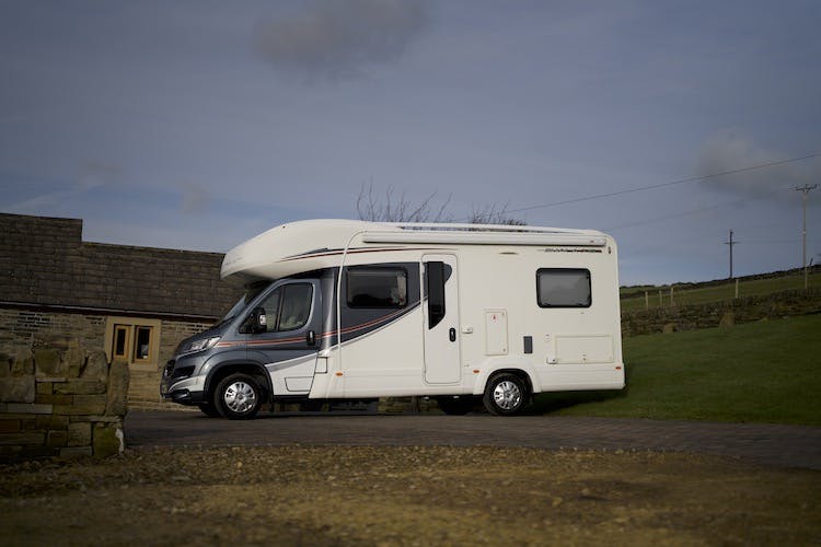 A 2014 Auto-Trail Imala 715 Lowline recreational vehicle is parked on a paved driveway. It has several windows and a door. The background includes a stone building and a grassy area with a fence. The sky is cloudy.