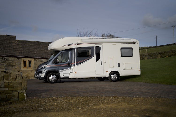 A 2014 Auto-Trail Imala 715 Lowline motorhome is parked on a cobblestone driveway next to a stone building with small windows. The surrounding area includes a grassy lawn, a stone wall, and a cloudy sky in the background.