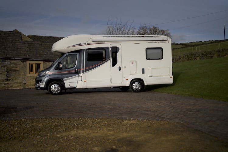 A white 2014 Auto-Trail Imala 715 Lowline motorhome with black and red accents is parked on a paved driveway next to a stone building. In the background, there is a grassy area with a few trees and hilltops visible under a partly cloudy sky.