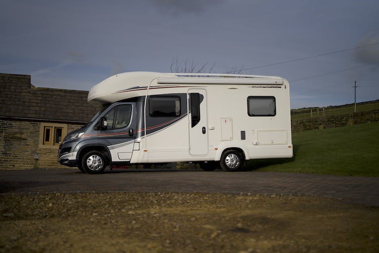 A white 2014 Auto-Trail Imala 715 Lowline camper van with black and red accents is parked on a paved driveway next to a brick building. The sky is overcast, and a grassy hill is visible in the background.