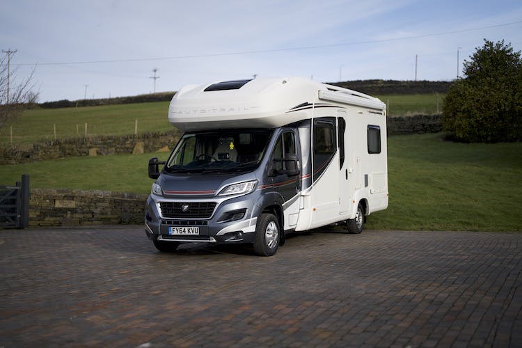 A white 2014 Auto-Trail Imala 715 Lowline motorhome is parked on a paved driveway. The area around the motorhome features a stone wall, grassy fields, and trees. The sky is clear with minimal cloud cover.