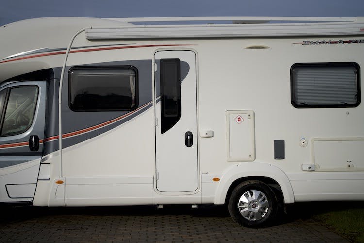 A white 2014 Auto-Trail Imala 715 Lowline motorhome is parked on a grey paved surface. The vehicle has a window near the front and another towards the rear, with a door in the middle. The body features red and grey stripes, and there is a small storage compartment next to the rear window.
