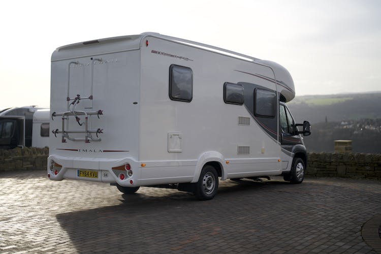 A white 2014 Auto-Trail Imala 715 Lowline camper van is parked on a paved surface with a scenic background. The van has a ladder on the back and a registration plate. It is clean and appears to be in good condition. In the background, there is another vehicle and a view of a hilly landscape.