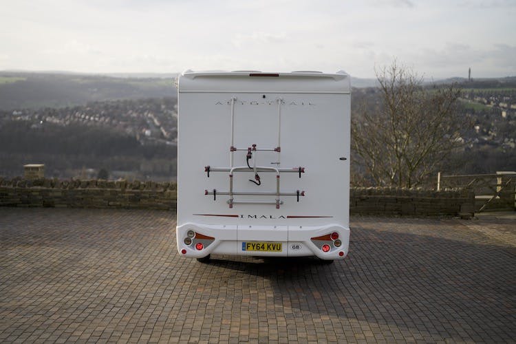 A white 2014 Auto-Trail Imala 715 Lowline motorhome, complete with a bike rack and a UK license plate, is parked on a paved area. The background features a hilly landscape with trees and distant buildings.