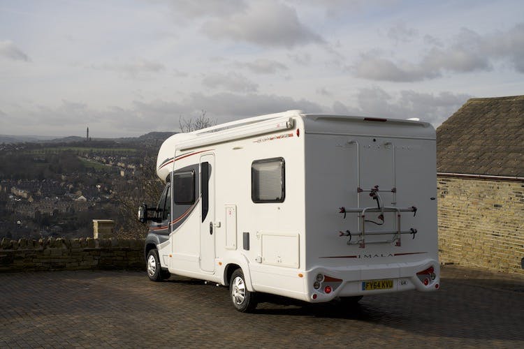 A white 2014 Auto-Trail Imala 715 Lowline motorhome is parked on a brick-paved area overlooking a town and countryside. The sky is partly cloudy. The motorhome, featuring a bike rack on the rear, appears stationary with its registration plate reading "FX64 KYA.