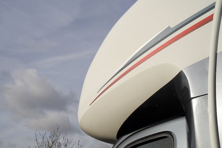 Close-up view of the upper section of a 2014 Auto-Trail Imala 715 Lowline against a cloudy sky. The vehicle features a streamlined design with red and gray stripe details near the roof. Minimal foliage is visible against the sky in the background.