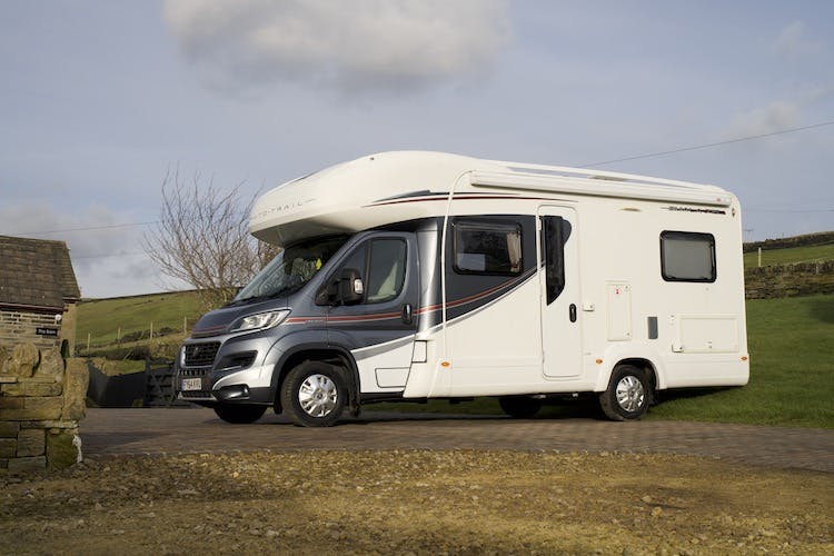 A 2014 Auto-Trail Imala 715 Lowline motorhome is parked on a paved driveway. The vehicle has a high roof area, large windows, and dark decorative stripes along the sides. The background features a stone building, grassy areas, and a cloudy sky.