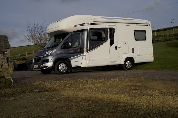 A 2014 Auto-Trail Imala 715 Lowline motorhome with grey and black accents is parked on a brick driveway. The background includes a grassy field, stone wall, and part of a wooden structure under a cloudy sky.