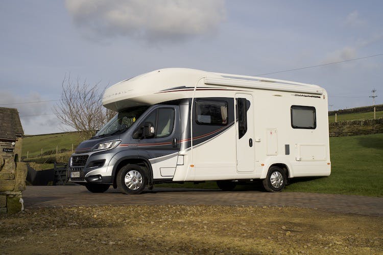 A 2014 Auto-Trail Imala 715 Lowline motorhome is parked on a paved area with a scenic, grassy backdrop. The vehicle has dark and red accents along the side and is situated in an outdoor rural setting with a partially visible building and stone wall on the left.