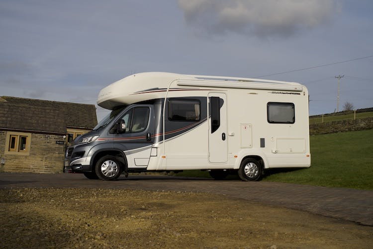 A 2014 Auto-Trail Imala 715 Lowline motorhome, featuring gray and red detailing and a large side window, is parked on a paved area next to a small building with stone walls. The surrounding area includes some grass and a cloudy sky.