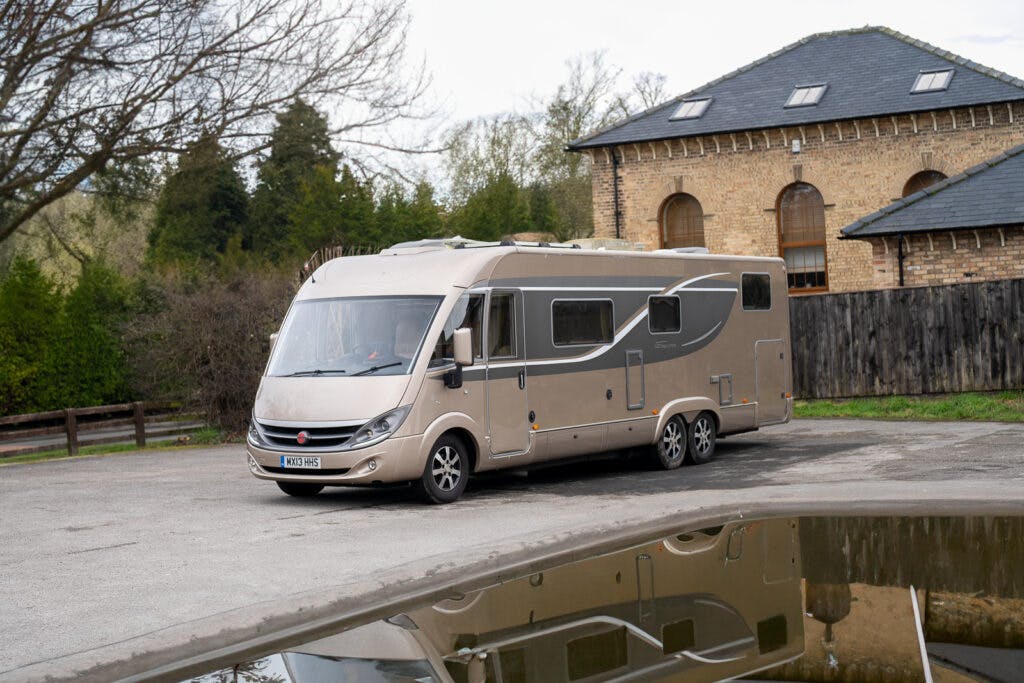 A large beige 2013 Burstner Elegance i810 G motorhome is parked in a lot beside a stone building with a slate roof. The vehicle has multiple windows and additional storage compartments. In the foreground, the reflective surface of water adds tranquility. Trees and a wooden fence are in the background.