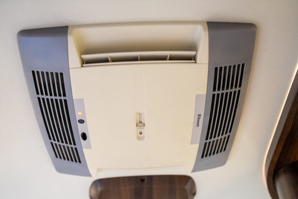 A ceiling-mounted air conditioning unit, perfect for a 2013 Burstner Elegance 810 G, features a beige center panel and gray side grilles. The vent in the middle has adjustable louvers. Control buttons and indicator lights are located on the left side of the panel. The brand name "Dometic" is visible.