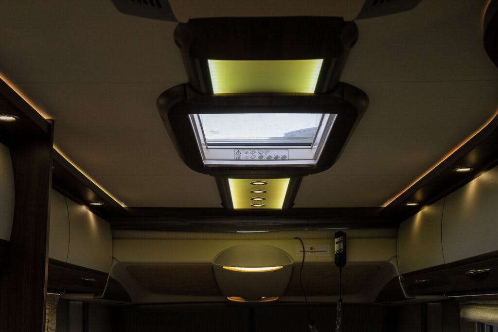 The image shows the interior ceiling of a 2013 Burstner Elegance 810 G, featuring recessed overhead lighting and a skylight window. The ceiling has wood paneling and overhead storage compartments on the sides. An electric control switch is visible near the skylight.